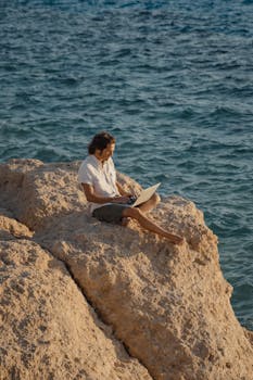 Man working on a laptop, sitting on a cliff overlooking the ocean, embodying the digital nomad lifestyle.
