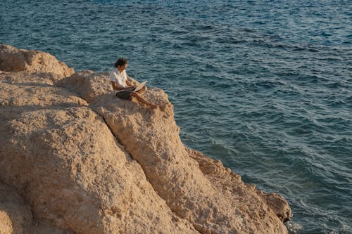 Man in White Sleeve Shirt Sitting on Brown Rock Formation Near Body of Water