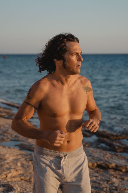 Free A man jogs shirtless along a beach at sunset, embodying fitness and freedom. Stock Photo