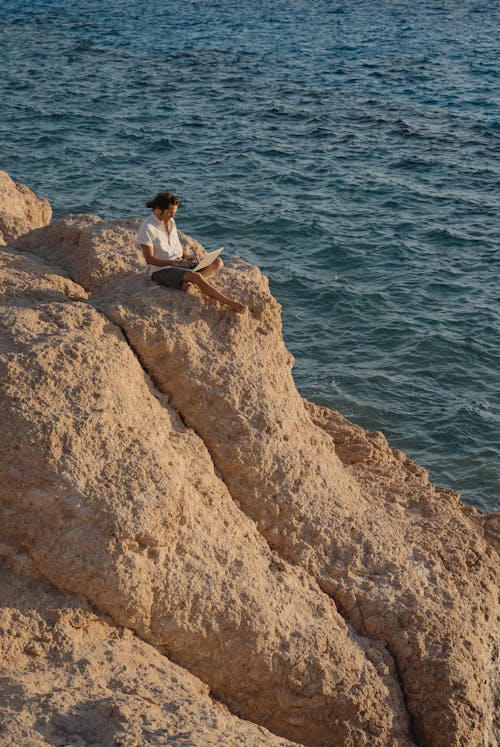 Man in White Dress Shirt Sitting on Brown Rock Formation Near Body of Water