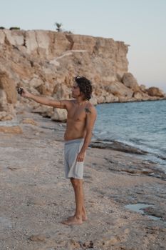 A shirtless man takes a selfie at a rocky beach at sunset, showcasing serene coastal vibes.