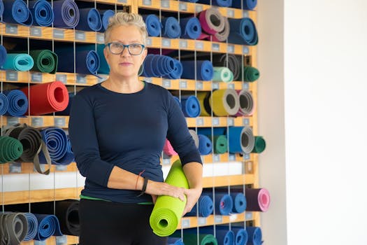 Mature woman standing with yoga mat in a well-organized studio. Bright and serene environment.