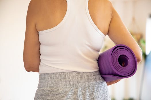 Woman in white tank top holding a rolled purple yoga mat indoors, preparing for exercise.