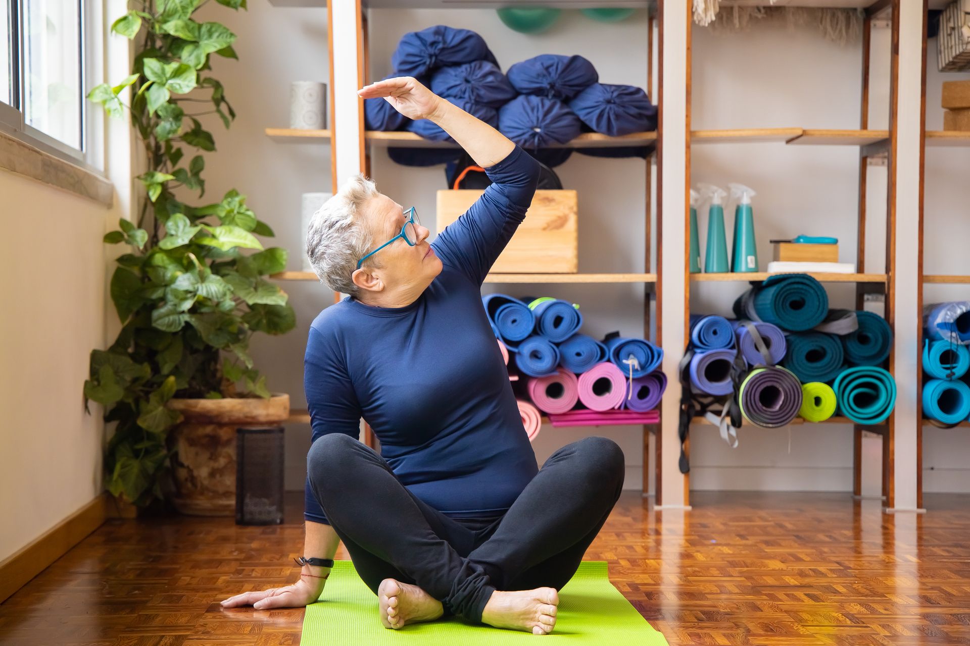 Elderly woman practicing yoga seated indoors on a green mat, promoting healthy lifestyle.