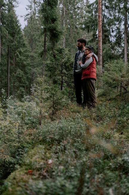 A couple stands embracing in a serene forest, surrounded by lush greenery and tall trees.
