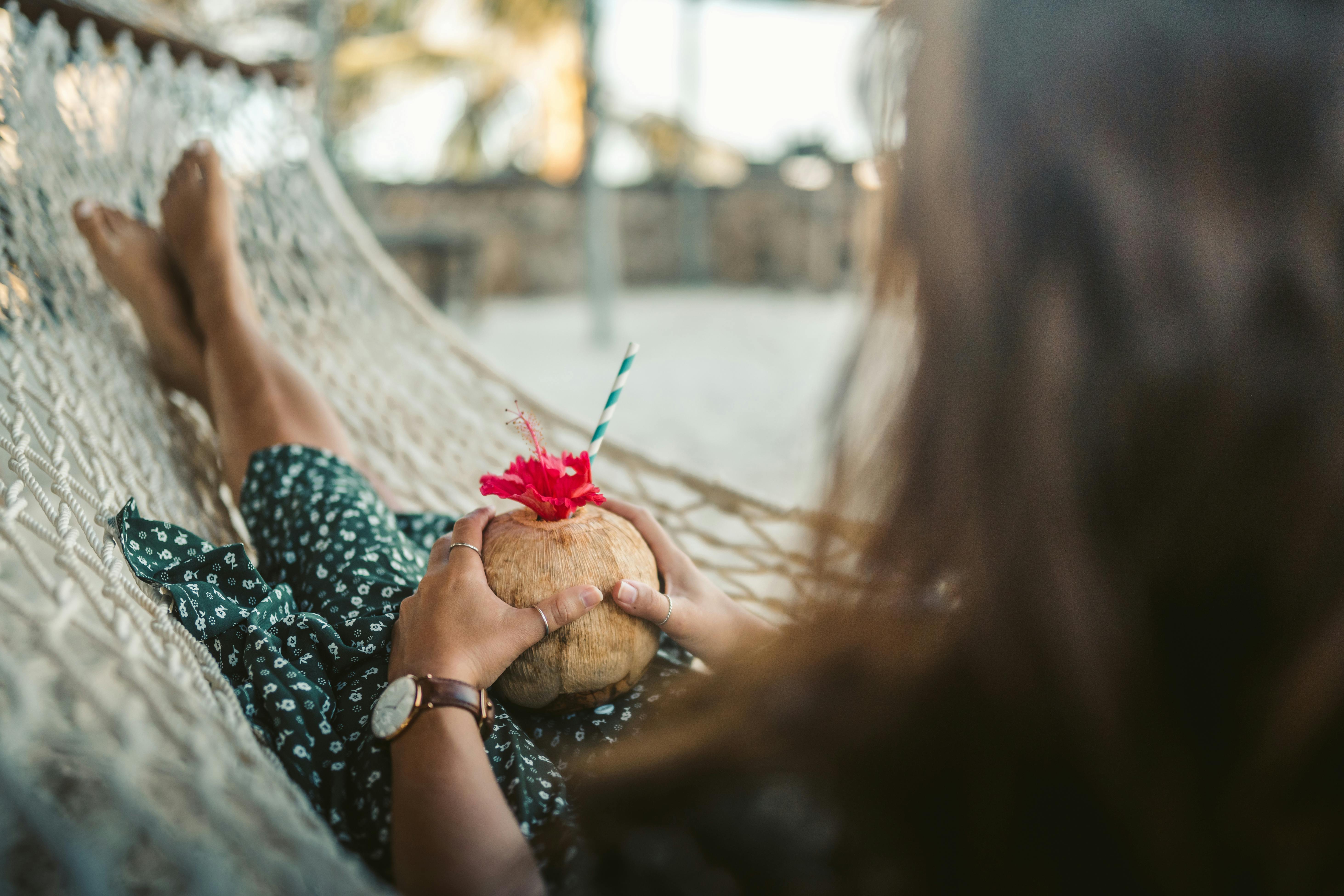 A woman enjoys a coconut drink while relaxing in a hammock on a sandy beach.