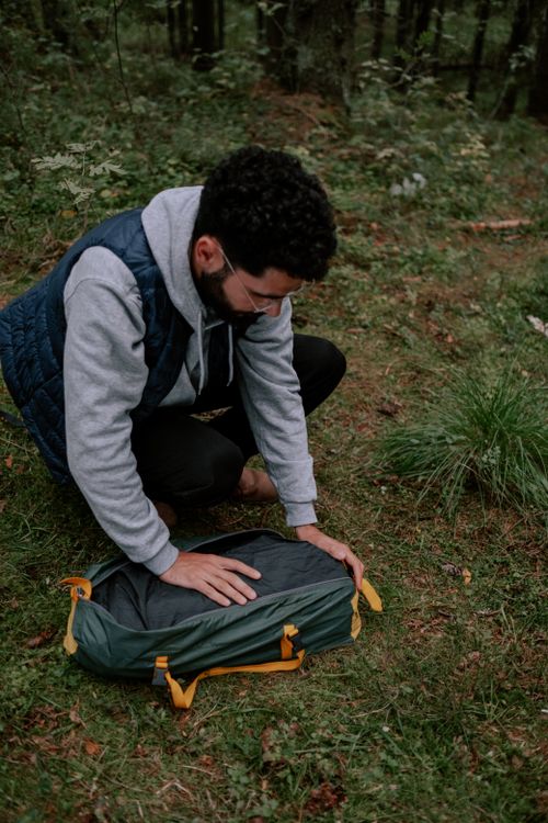 Free A man in a hoodie and vest organizes camping gear in a forest setting. Stock Photo