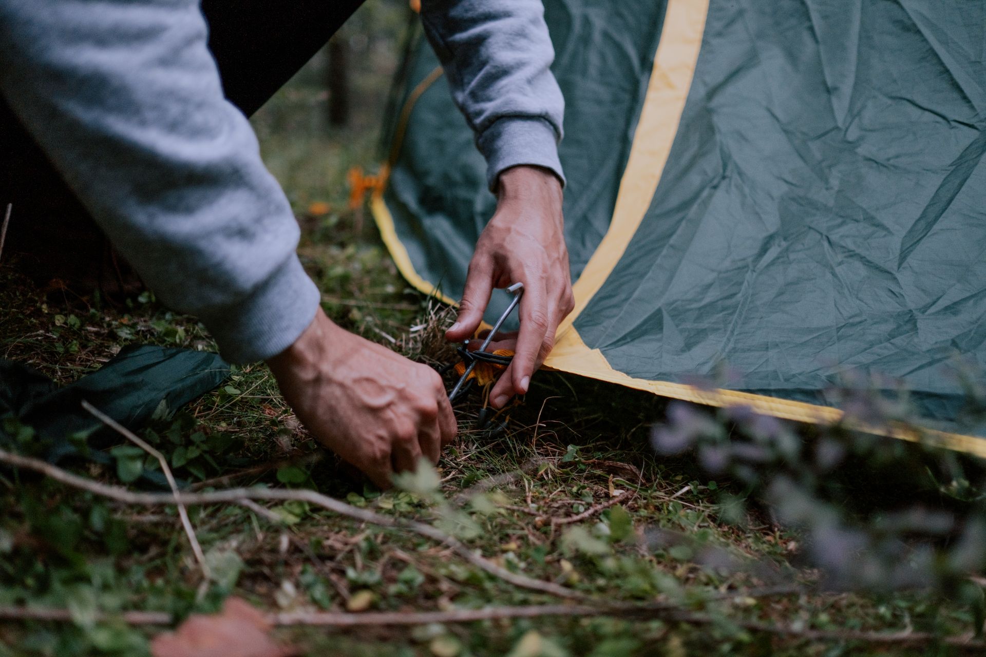 A close-up of a person securing a tent peg in a forested campsite, depicting outdoor camping activit
