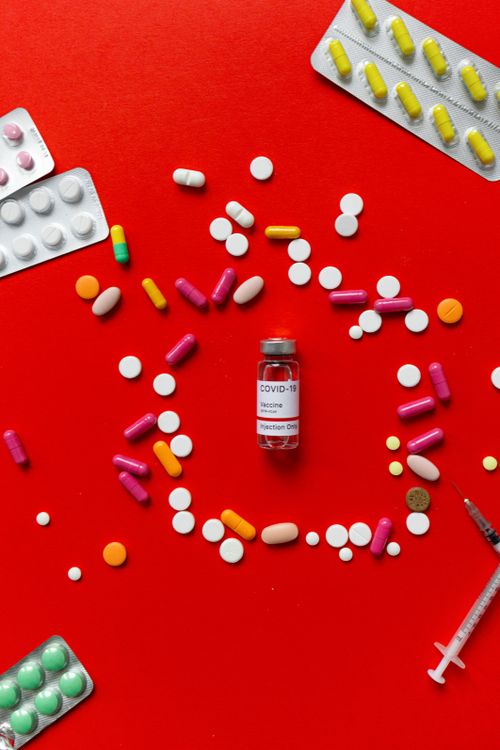 Free Flat lay of various pills and COVID-19 vaccine vial on a vibrant red surface. Stock Photo
