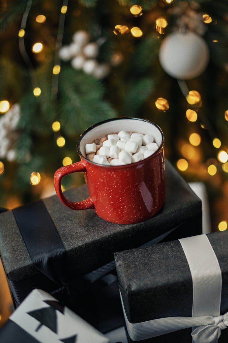 Hot Chocolate With Marshmellows Standing In Front Of A Christmas Tree On Boxes With Presents