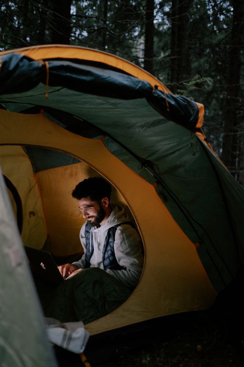 Free A bearded man relaxes in a tent using a laptop during an evening camping trip. Stock Photo