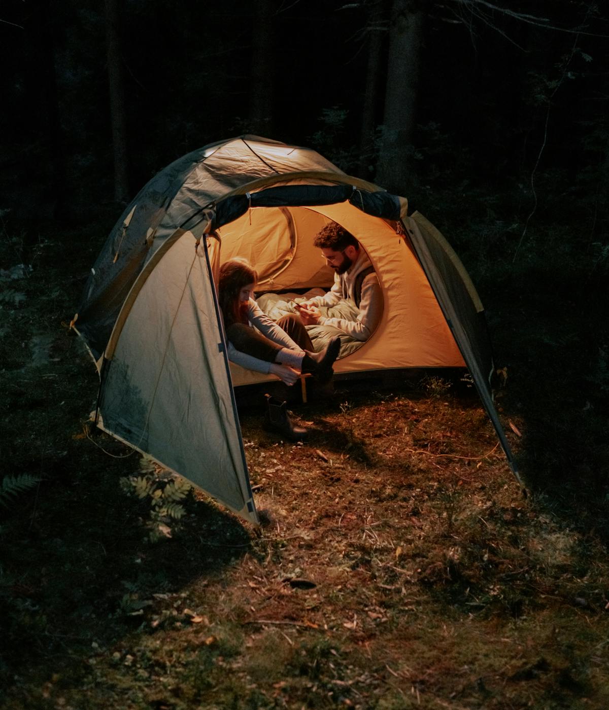 The image shows a couple sitting inside an illuminated camping tent in a dark, wooded area.
