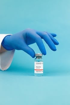 A hand in blue gloves handling a COVID-19 vaccine vial against a blue background.