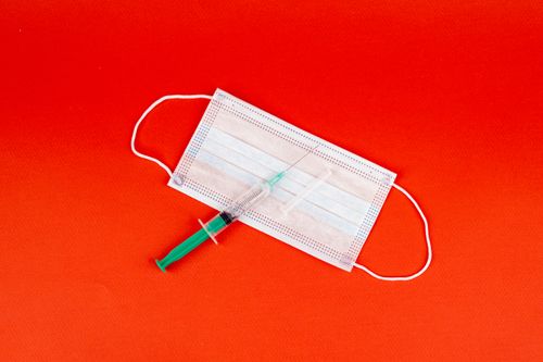 Free A syringe placed on a face mask against a vivid red surface, symbolizing healthcare. Stock Photo