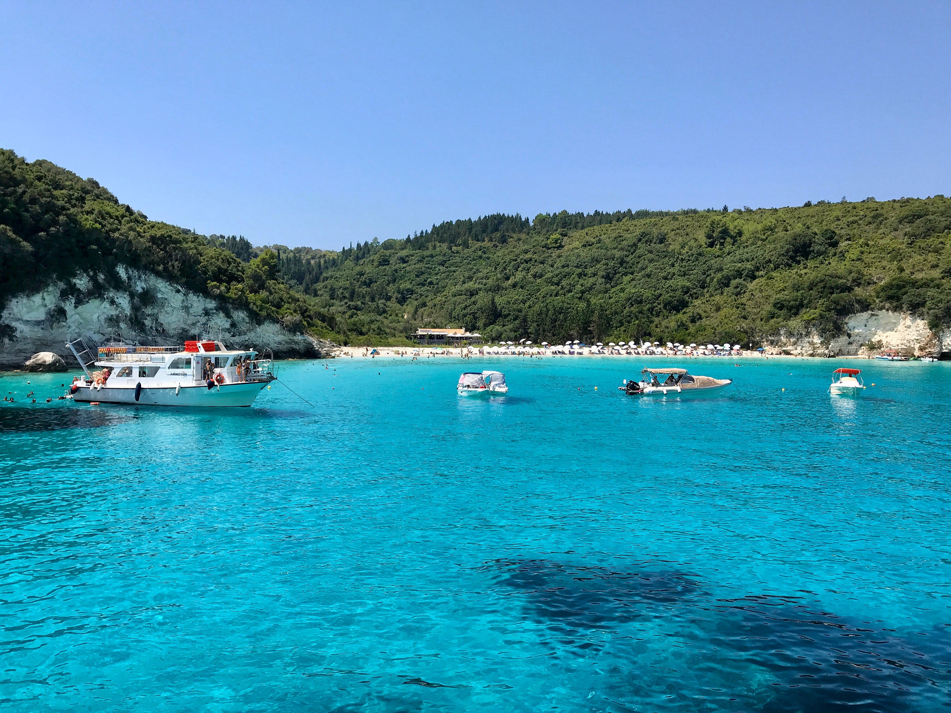 Beautiful turquoise waters with boats near lush green Antipaxos island, Greece.