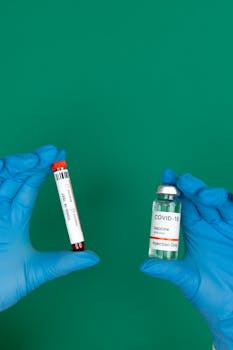 Close-up of hands in gloves holding a blood sample and COVID-19 vaccine vial with green background.