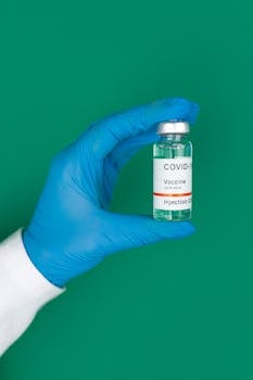 A medical professional's gloved hand holding a COVID-19 vaccine vial against a green background.