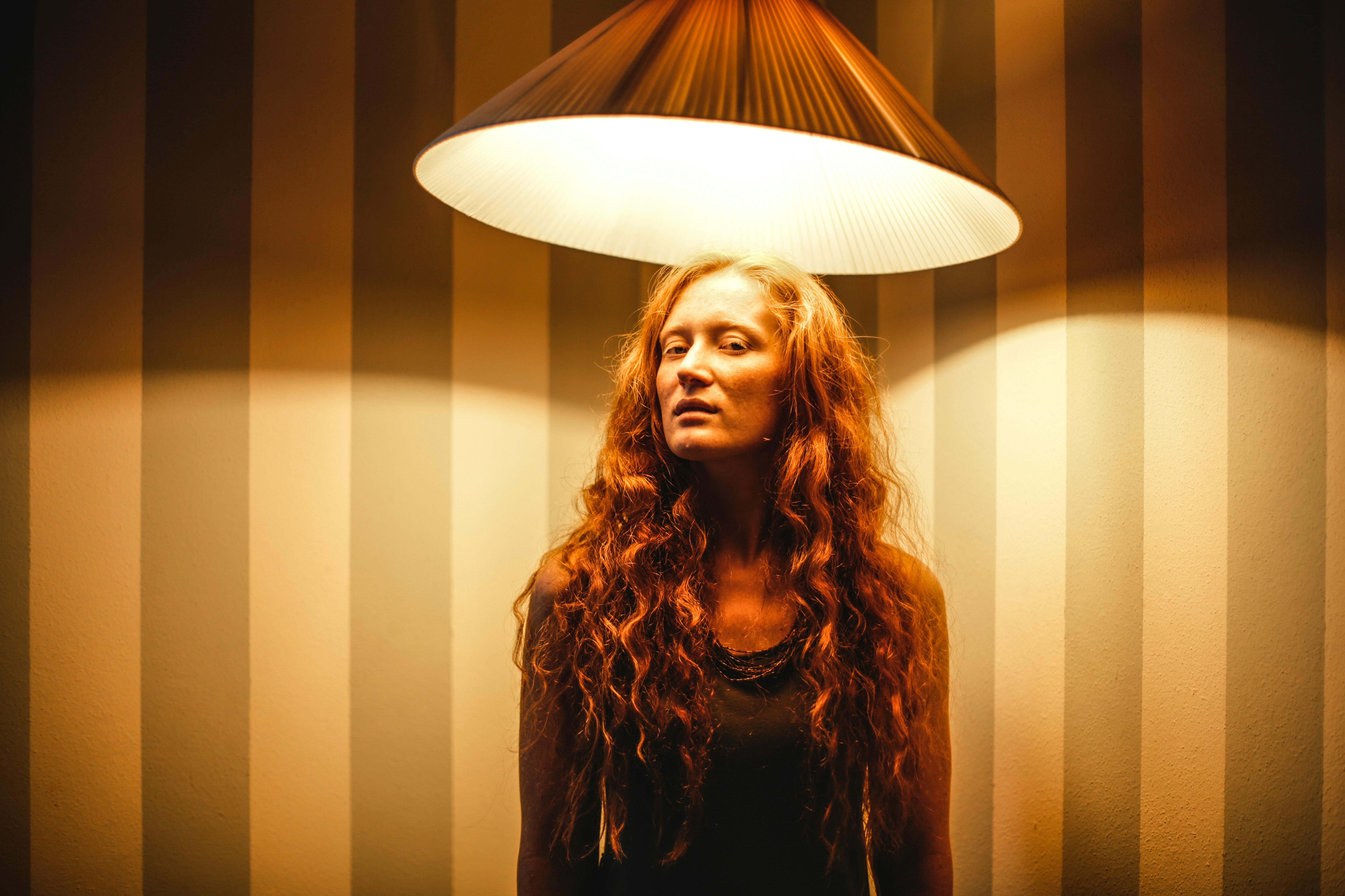 Serious female with red curly hair looking at camera while standing under glowing lamp illuminating in dark room with shadows on striped wall