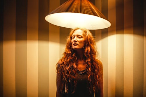 Serious female with red curly hair looking at camera while standing under glowing lamp illuminating in dark room with shadows on striped wall