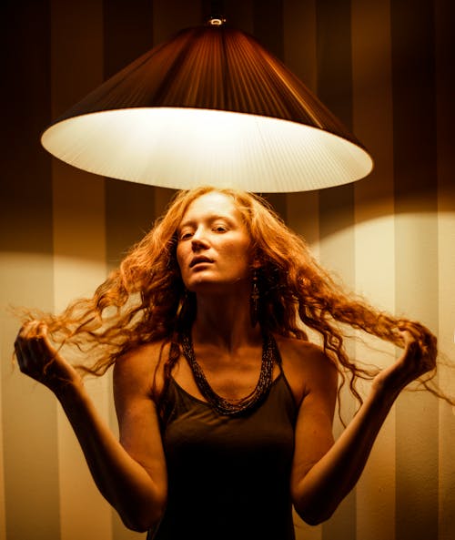 Attractive female looking at camera and touching red hair while standing under glowing lamp against striped wall at low light