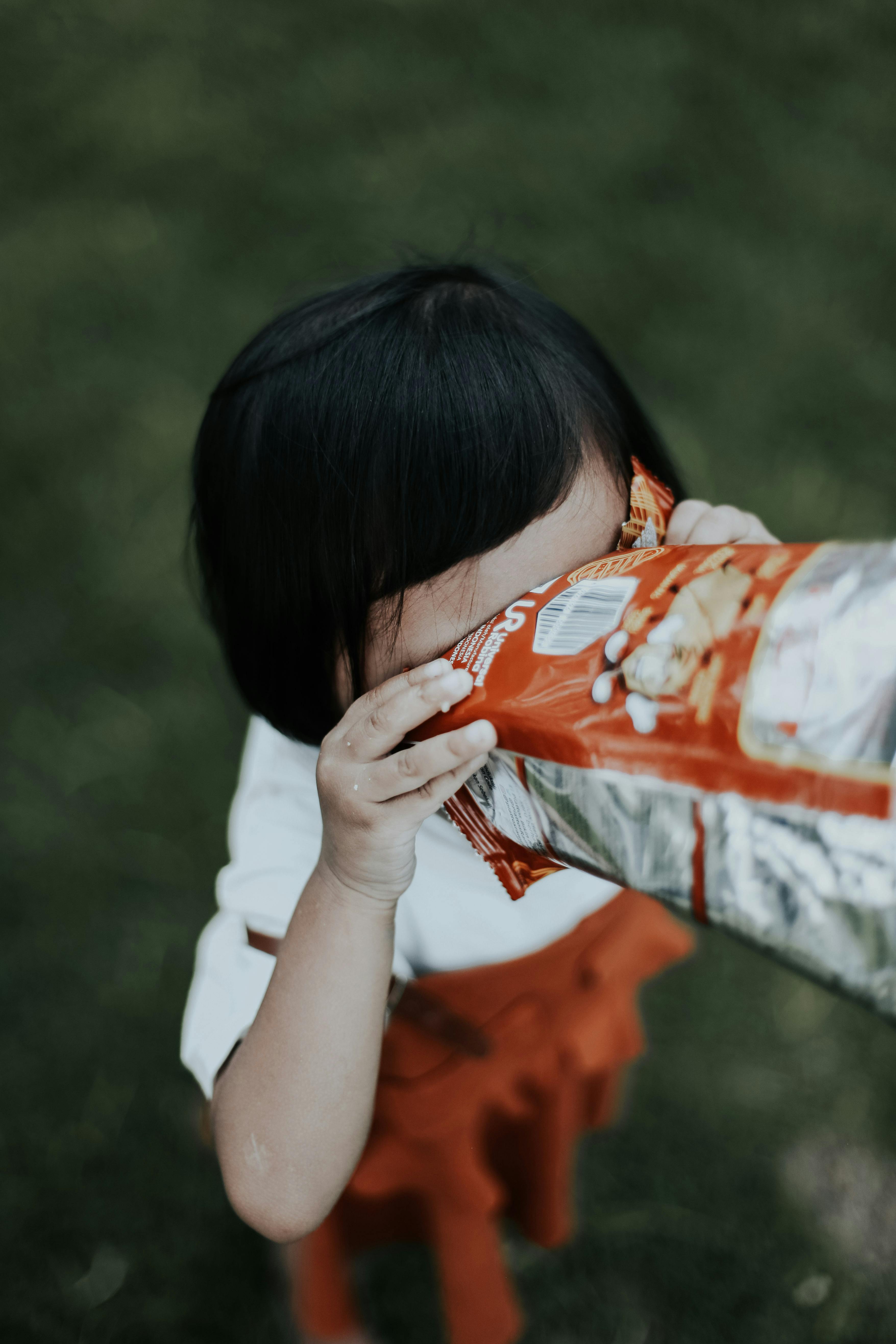 A young girl eagerly eats chips from a bag in a vibrant outdoor setting.