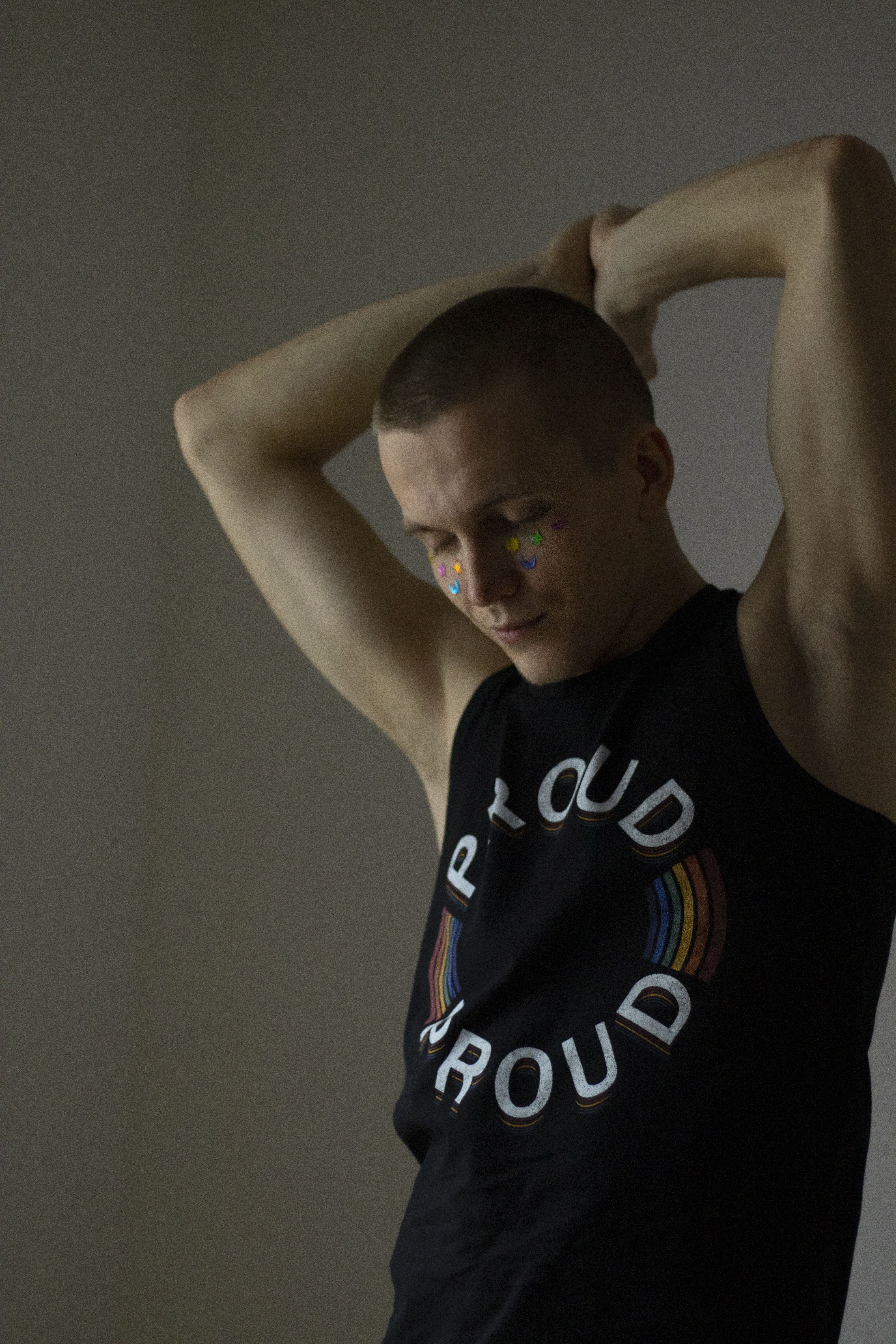 A man confidently posing with raised arms in a black tank top labeled 'Proud' in a studio setting.