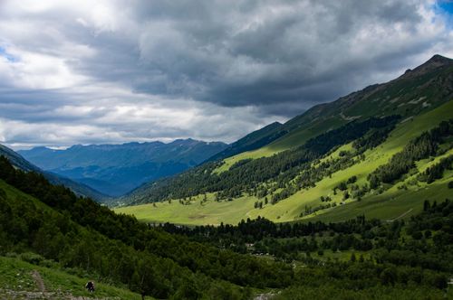 Free Explore a scenic mountain valley with rolling green hills, lush forests, and dramatic cloud formations. Stock Photo