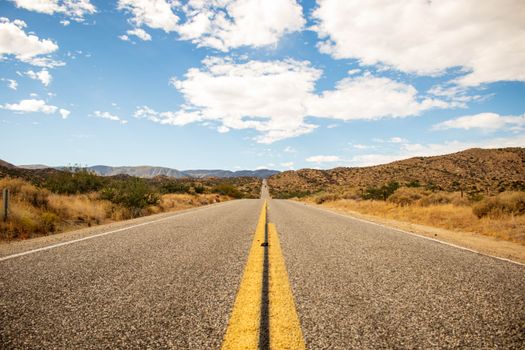 Desert highway stretching into mountainous horizon under bright blue sky and clouds.