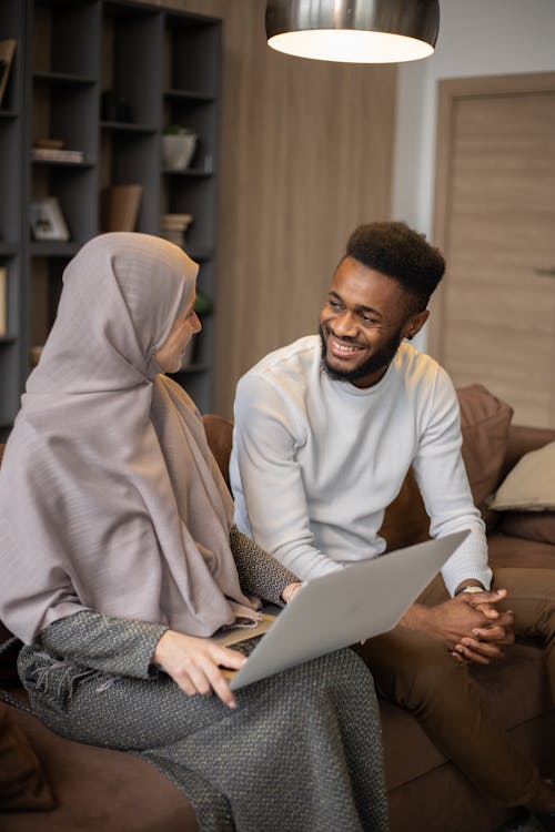 Positive young multiracial couple in casual outfits sitting on sofa at home with laptop and looking at each other