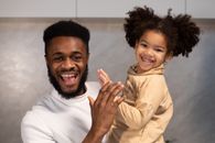 Positive young African American father with beard in casual clothes carrying smiling daughter and looking at camera