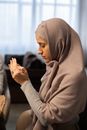 Side view of thoughtful young Muslim woman in hijab sitting on chair at table and praying before eating at home