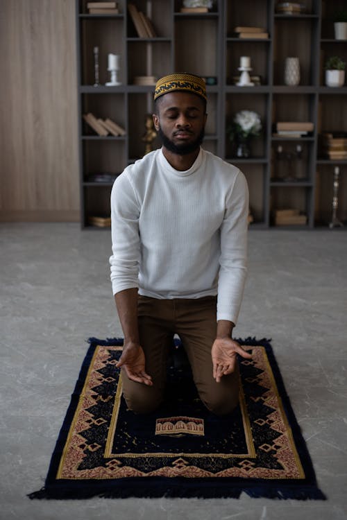 Full body concentrated young African American male with closed eyes in hat praying on rug at home