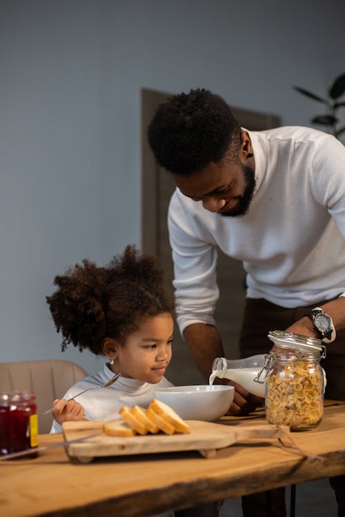 Happy young African American father pouring milk into bowl for daughter for eating cornflakes while having breakfast together at home