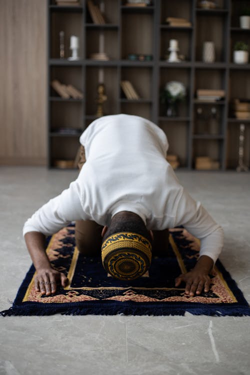 Focused anonymous Muslim African American male in hat praying on rug on floor at home