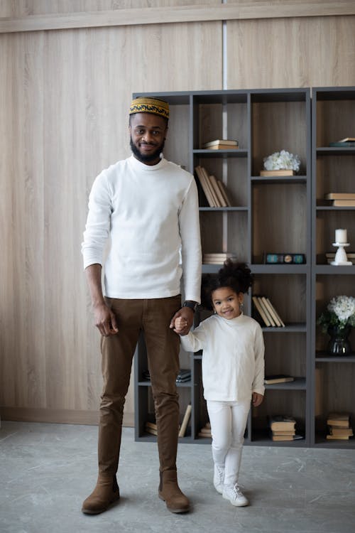 Full body positive bearded African American father in hat holding hands with cute little daughter looking at camera