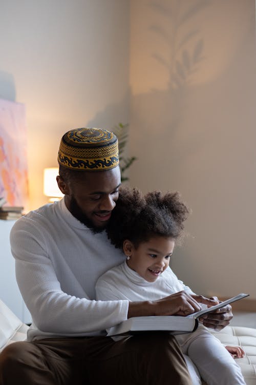 Free Delighted black male in turban and smiling daughter sitting in light room and reading book while having rest Stock Photo