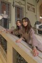 Unemotional young females in stylish jackets leaning on marble stairs railing and looking at camera while spending time in grand gallery