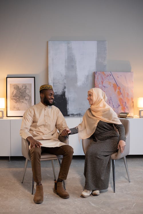 Happy Muslim woman and black male in traditional clothes looking at each other and holding hands