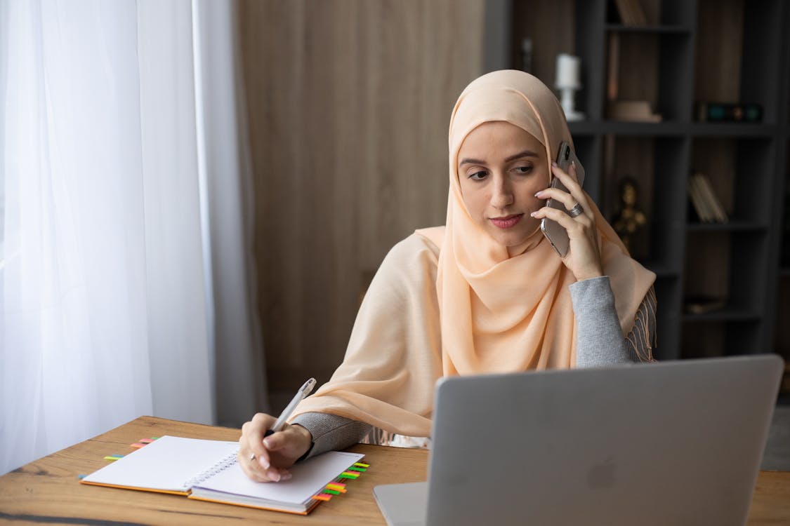  A Muslim woman wearing a hijab is working on her laptop and taking notes while talking on the phone.