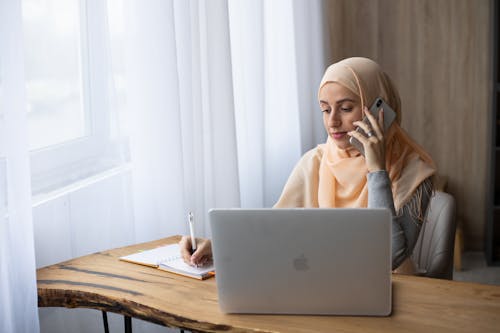 Free Concentrated Muslim female freelancer writing notes in notebook and having conversation on mobile phone while working Stock Photo