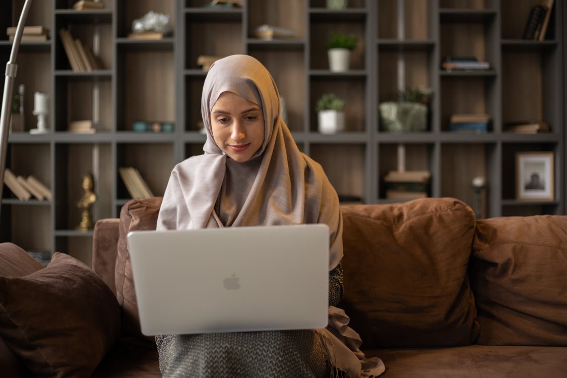 Pensive muslim female remote worker sitting on comfortable sofa and working on project on laptop