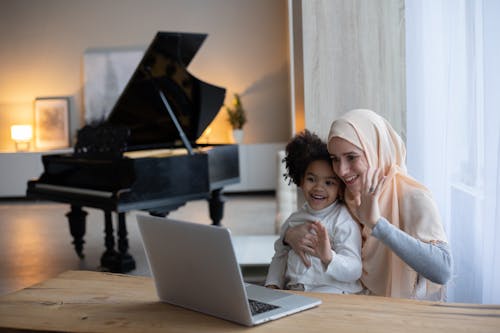 Free Positive Muslim mother and adorable African American girl sitting at table near window and having video call on portable laptop Stock Photo