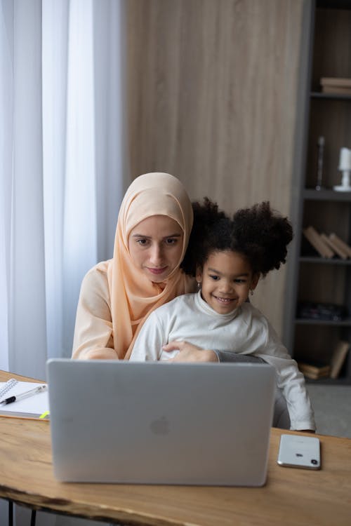 Concentrated Muslim woman and black daughter sitting at table with laptop