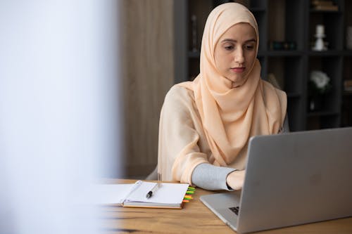 Focused young Muslim woman wearing elegant headscarf sitting at wooden table with planner and using modern laptop while working remotely