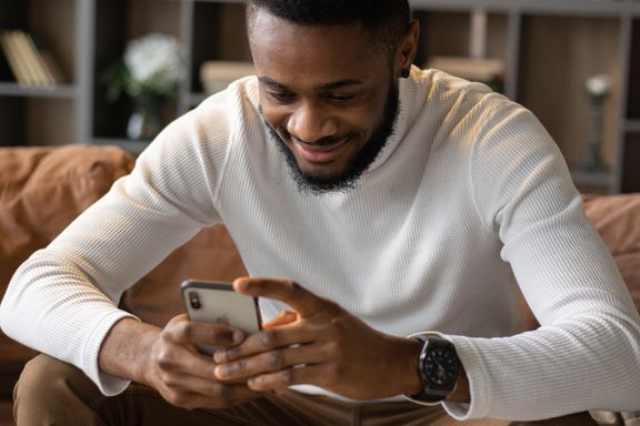 A man seated on a brown leather sofa smiles softly while looking at his smartphone