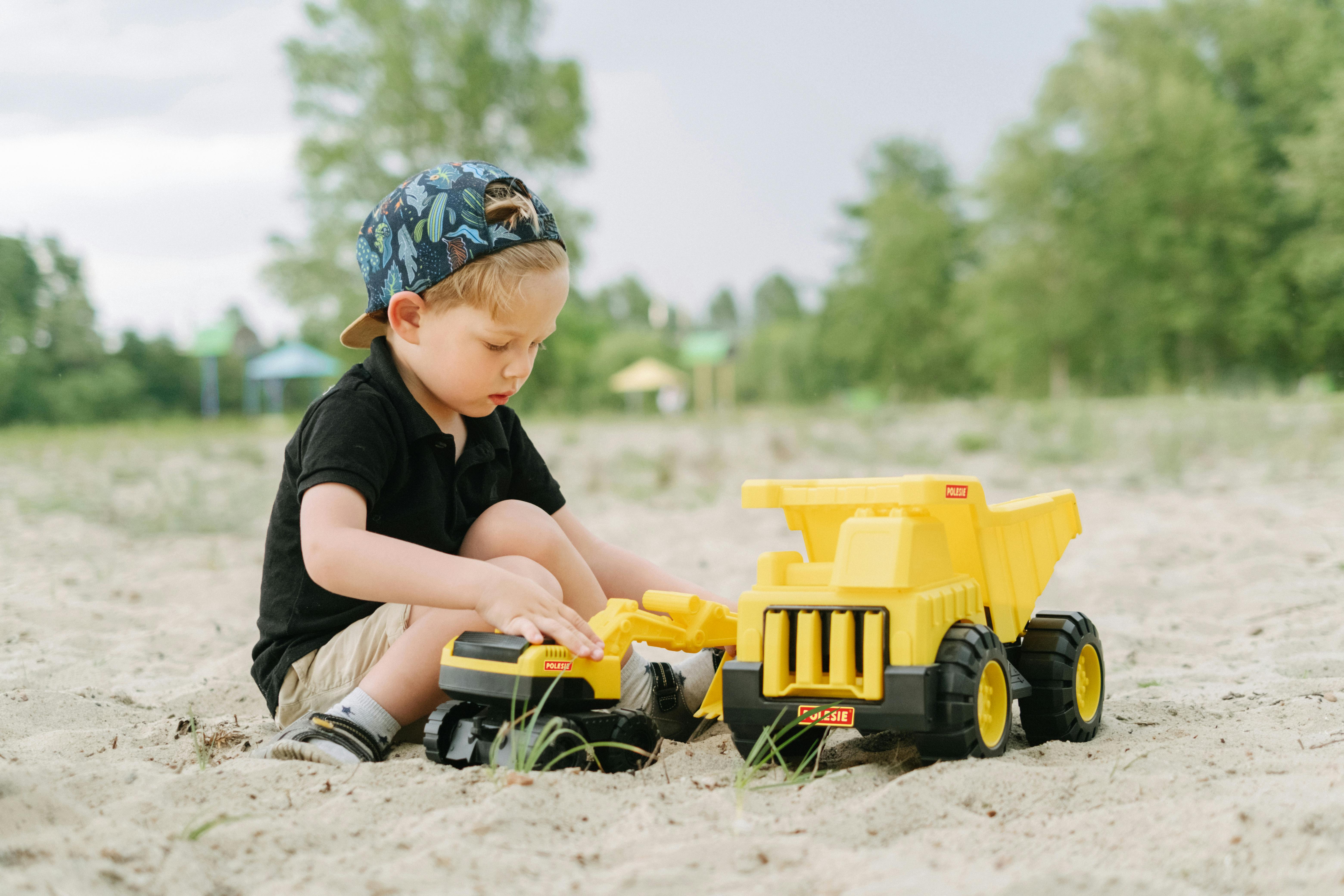 A young boy plays outdoors with a yellow toy truck in the sand, enjoying summertime fun.