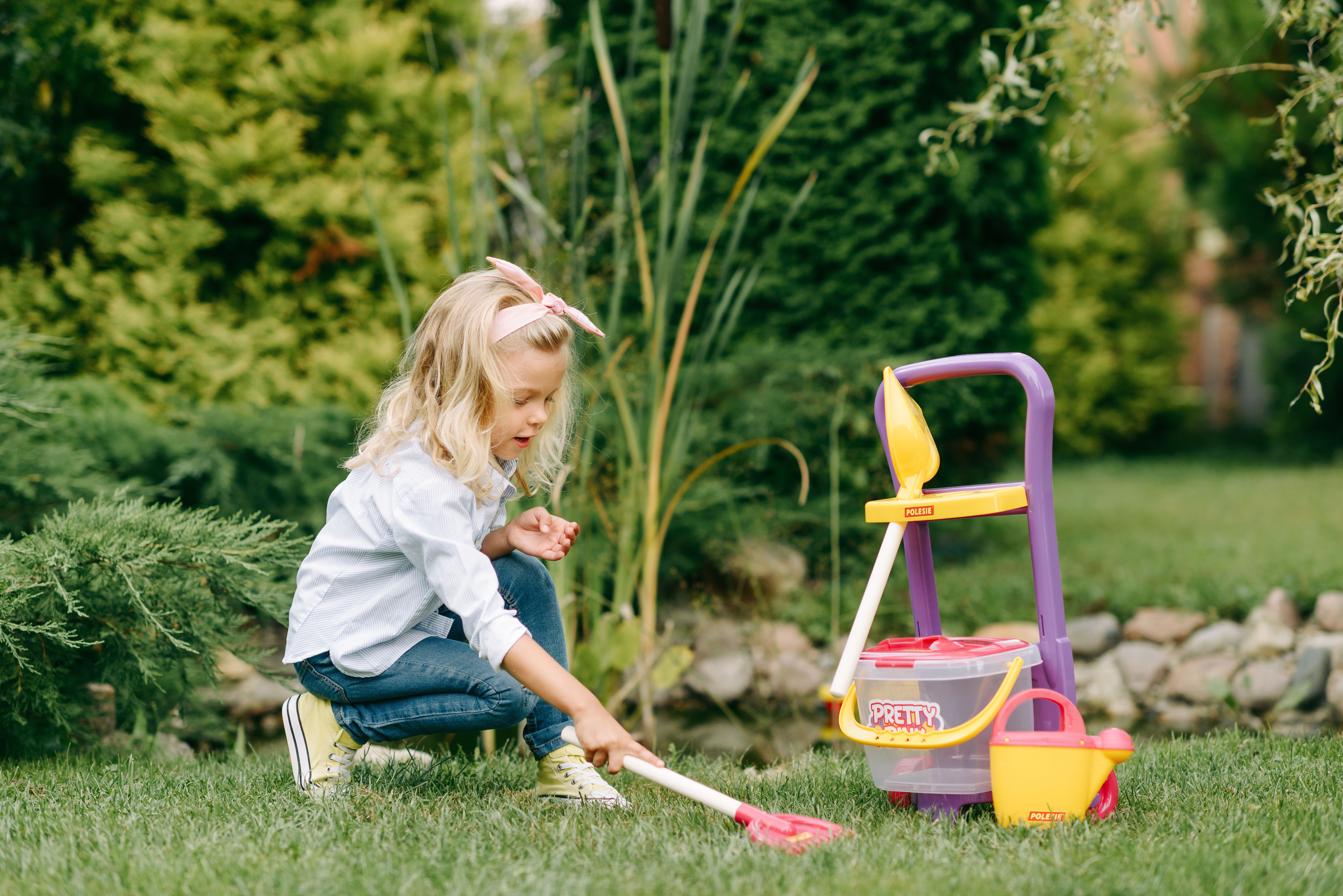 Blond girl playing with cleaning toys in an outdoor garden setting.