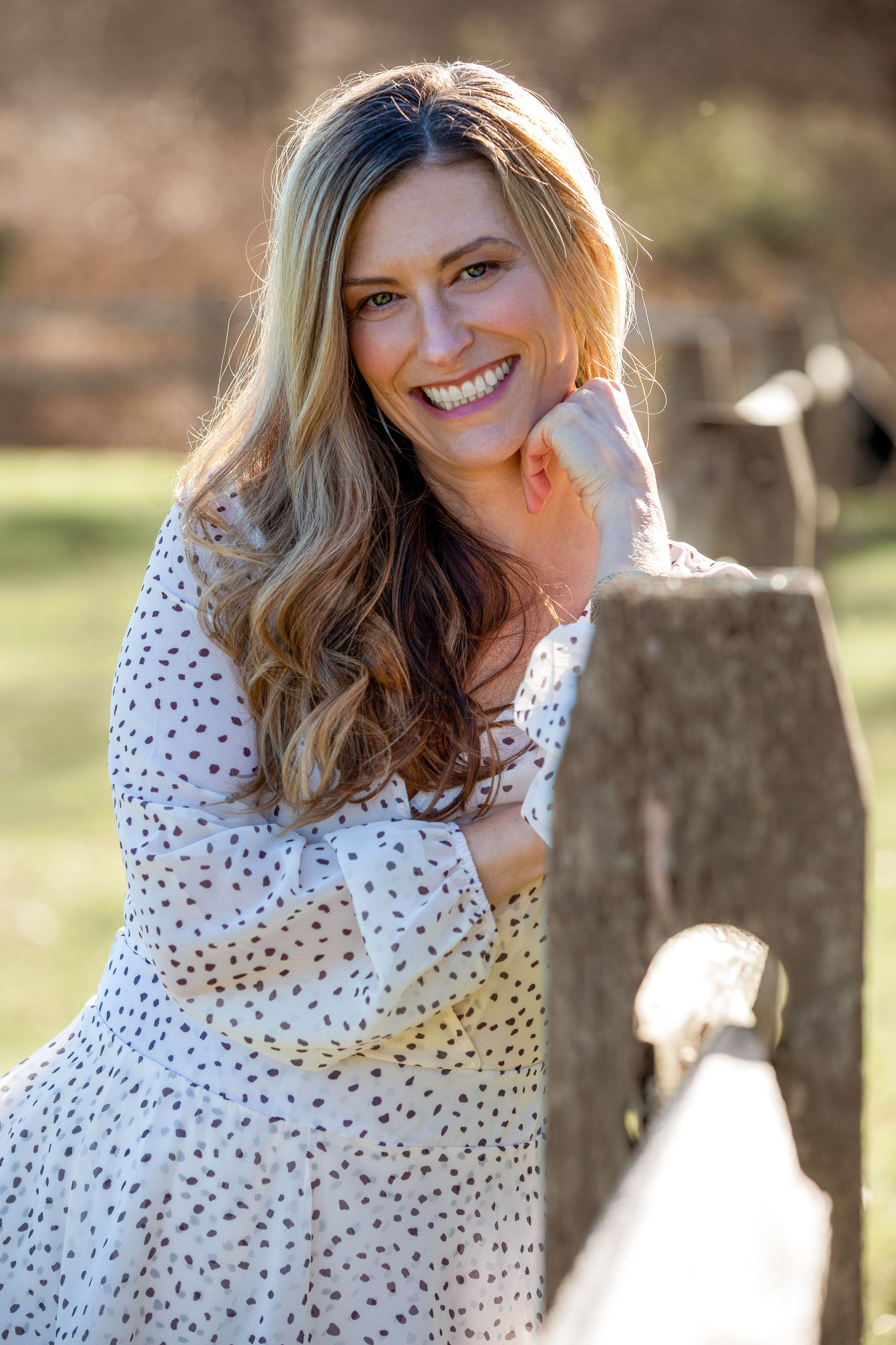 Cheerful female wearing polka dot dress looking at camera while standing at wooden fence in sunny countryside on blurred background