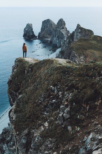 A solitary hiker in yellow stands on a dramatic cliff, overlooking rugged sea rocks.