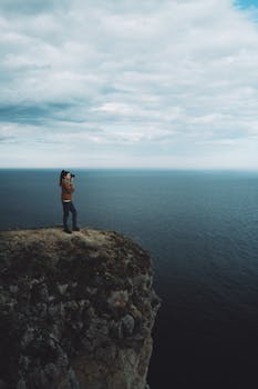 A woman stands on a cliff overlooking the vast ocean, capturing the scene with her camera.